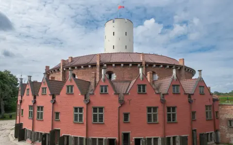 Okazały ceglany budynek z wieżą i czerwonymi domkami w kształcie podkowy.