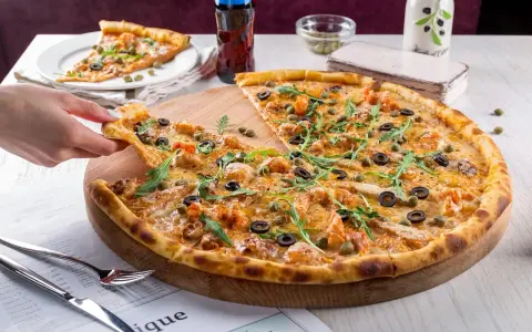 Pizza z krewetkami, oliwkami i kaparami na drewnianej desce, ręka podnosi kawałek.
