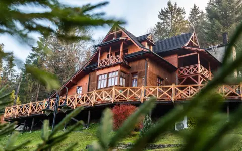 Drewniany góralski pensjonat z tarasem i balustradą, otoczony drzewami.