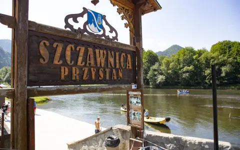 Drewniany szyld "Szczawnica Przystań" nad rzeką z tratwami i górami w tle.