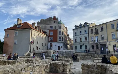 Ruiny zamku w Lublinie z ludźmi odpoczywającymi na kamiennych murach i zabytkowymi kamienicami w tle.