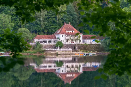 Dom z czerwonym dachem nad jeziorem, odbijający się w wodzie, otoczony zielenią.