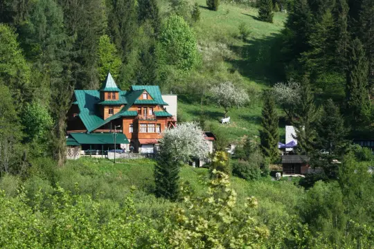 Drewniany schronisko PTTK z zielonym dachem otoczone lasem i zielonymi wzgórzami.