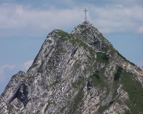 Giewont z krzyżem na szczycie, skaliste zbocza, zielona trawa, błękitne niebo z chmurami.