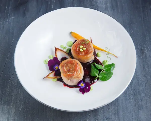 Eleganckie danie z dwóch medalionów mięsa zawiniętych w boczek, podane z purée, marchewką i kwiatami.