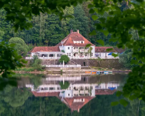 Dom z czerwonym dachem nad jeziorem, odbijający się w wodzie, otoczony zielenią.