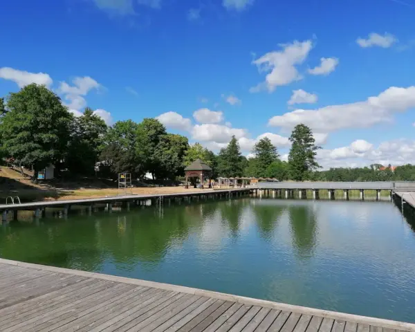 Drewniany pomost nad zieloną wodą jeziora Czos, z altaną i drzewami w tle, pod błękitnym niebem z chmurami.