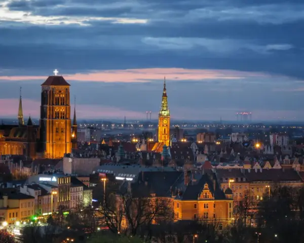 Widok na oświetlone zabytkowe centrum Gdańska o zmierzchu, z dominującą Bazyliką Mariacką i Ratuszem Głównego Miasta.