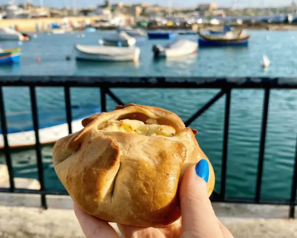 Ręka trzymająca pieczone ciastko z serem na tle maltańskiego portu z łodziami.