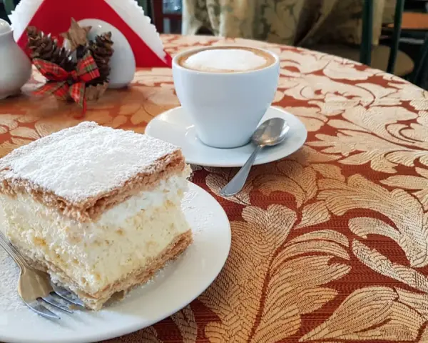 Kremówka posypana cukrem pudrem i filiżanka kawy na stoliku.