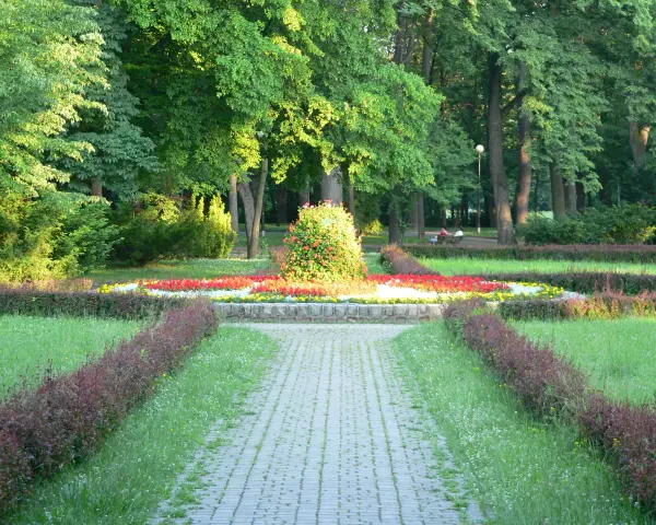 Kamienista ścieżka prowadzi przez park z kwitnącymi rabatami kwiatowymi i zielenią.