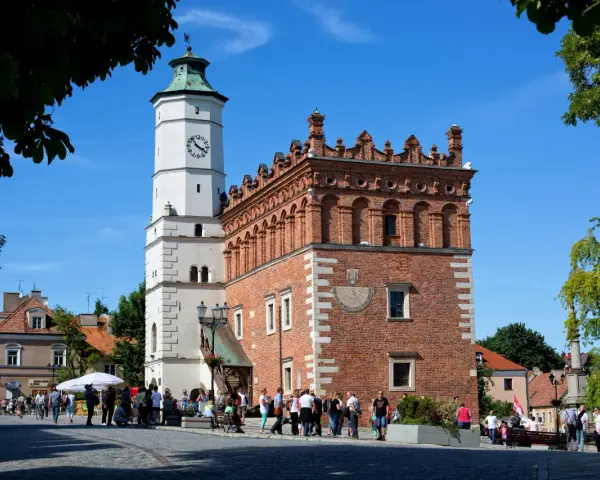 Ratusz w Sandomierzu z zegarem i zegarem słonecznym, na tle błękitnego nieba i drzew.