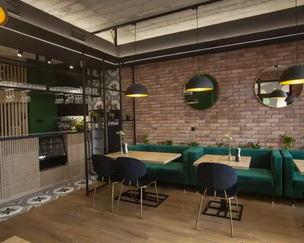 Nowoczesne wnętrze restauracji z ceglaną ścianą, zielonymi kanapami i drewnianymi stołami.
