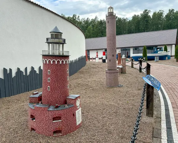 Miniatury latarni morskich na żwirowym placu, z budynkiem i łodzią w tle.
