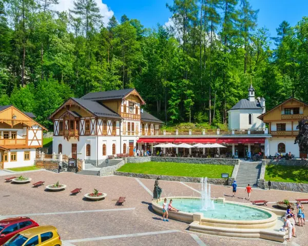 Plac Dietla w Szczawnicy z fontanną, budynkami uzdrowiskowymi i lasem w tle.