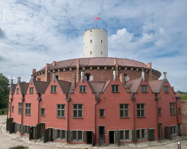 Okazały ceglany budynek z wieżą i czerwonymi domkami w kształcie podkowy.