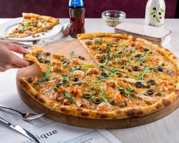 Pizza z krewetkami, oliwkami i kaparami na drewnianej desce, ręka podnosi kawałek.