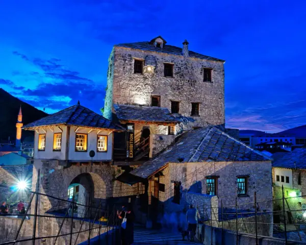 Kamienne budynki i most w Mostarze o zmierzchu, z widocznym minaretem w tle.