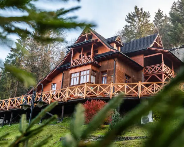 Drewniany góralski pensjonat z tarasem i balustradą, otoczony drzewami.