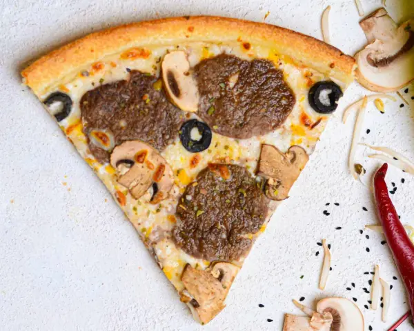 Kawałek pizzy z mięsem mielonym, pieczarkami i oliwkami na jasnym tle.