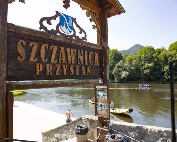Drewniany szyld "Szczawnica Przystań" nad rzeką z tratwami i górami w tle.