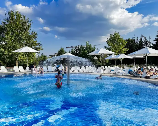 Rodzinna zabawa w hotelu z basenem na Mazurach. Dzieci pluskają się w wodzie, dorośli odpoczywają na leżakach pod białymi parasolami.
