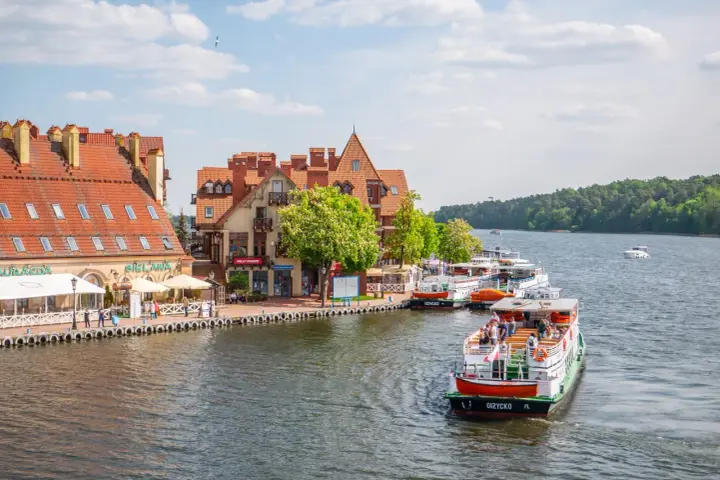 Port w Mikołajkach z zacumowanymi statkami wycieczkowymi i budynkami z czerwonymi dachami. Wspaniałe atrakcje w Mikołajkach czekają na turystów.