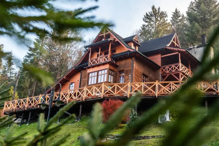 Drewniany góralski pensjonat z tarasem i balustradą, otoczony drzewami.