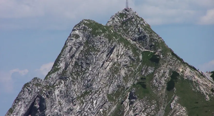 Giewont z krzyżem na szczycie, skaliste zbocza, zielona trawa, błękitne niebo z chmurami.