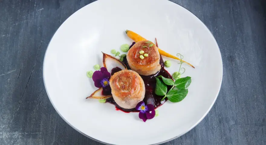 Eleganckie danie z dwóch medalionów mięsa zawiniętych w boczek, podane z purée, marchewką i kwiatami.