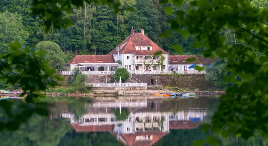 Dom z czerwonym dachem nad jeziorem, odbijający się w wodzie, otoczony zielenią.