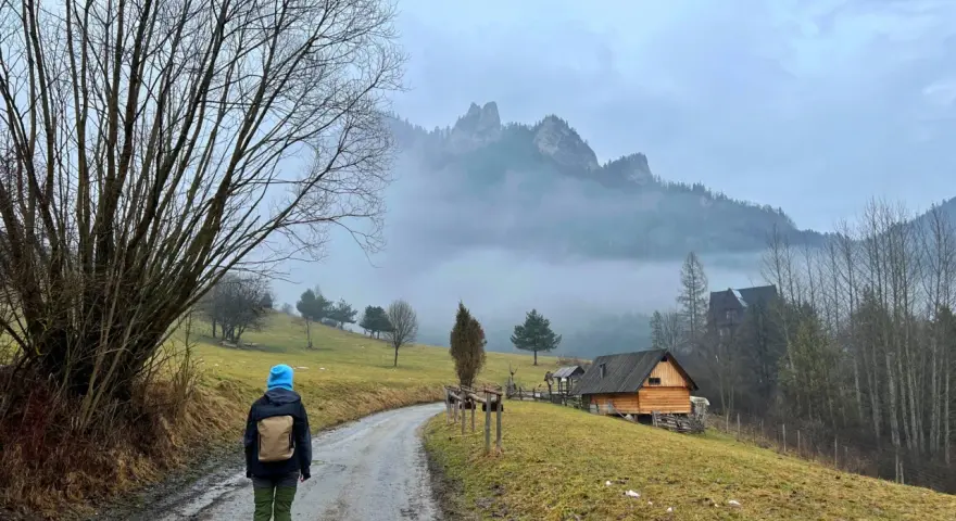Osoba w niebieskiej czapce idzie drogą w Pieninach, w tle góry i drewniane chatki.