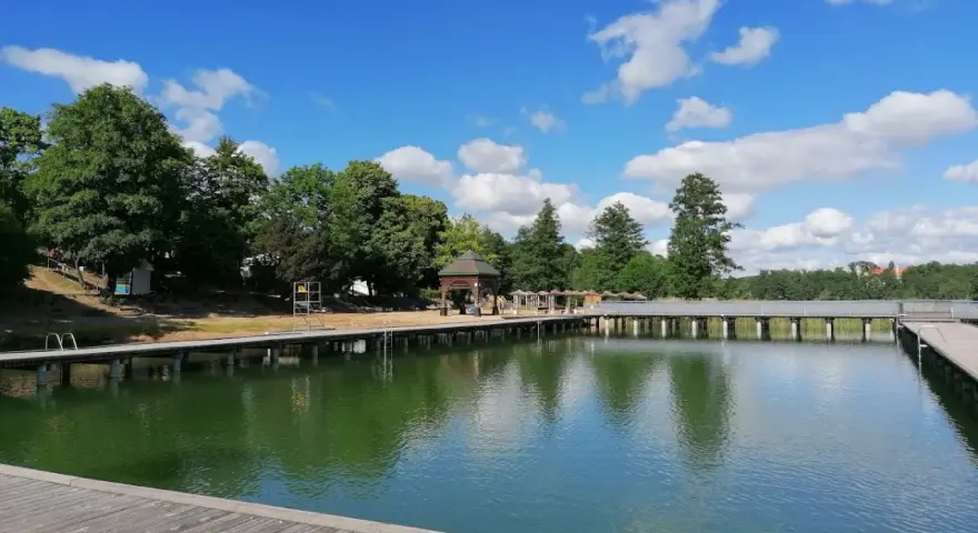 Drewniany pomost nad zieloną wodą jeziora Czos, z altaną i drzewami w tle, pod błękitnym niebem z chmurami.