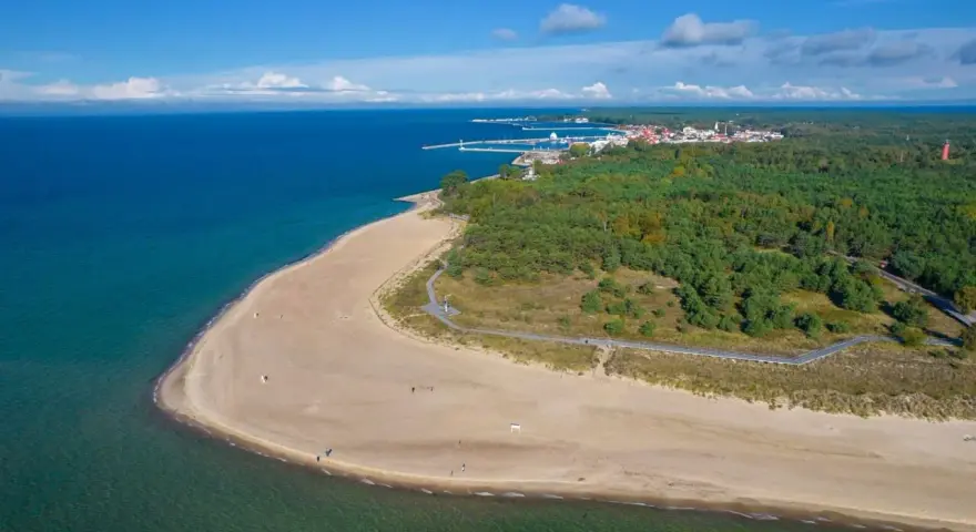 Widok z lotu ptaka na piaszczystą plażę, turkusowe morze i zielony las z miasteczkiem w tle.