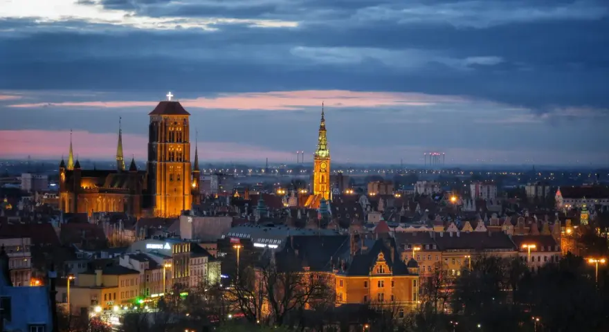 Widok na oświetlone zabytkowe centrum Gdańska o zmierzchu, z dominującą Bazyliką Mariacką i Ratuszem Głównego Miasta.