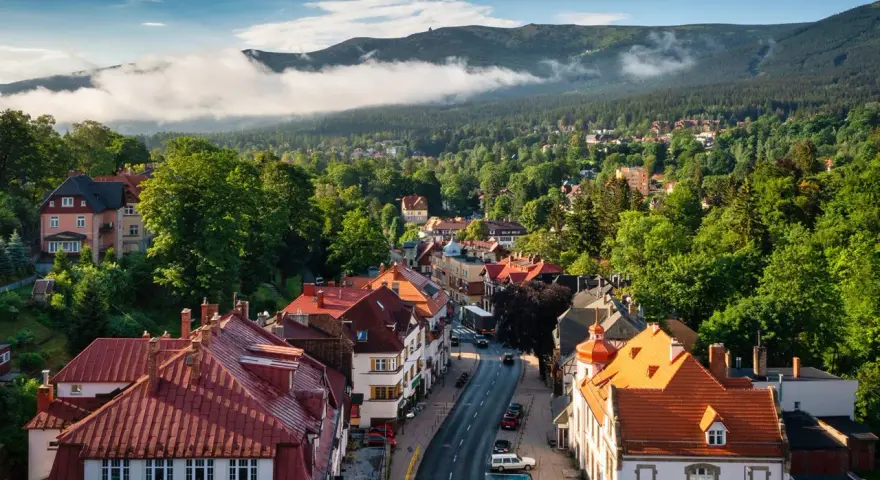 Widok z góry na miasteczko z czerwonymi dachami, otoczone zielonymi lasami i górami w tle.