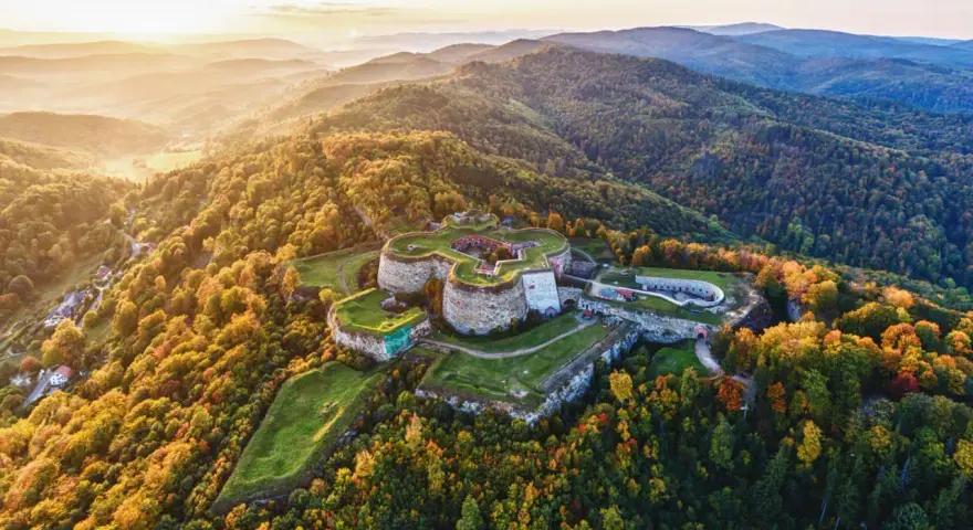 Twierdza Srebrna Góra otoczona jesiennym lasem, w świetle zachodzącego słońca.