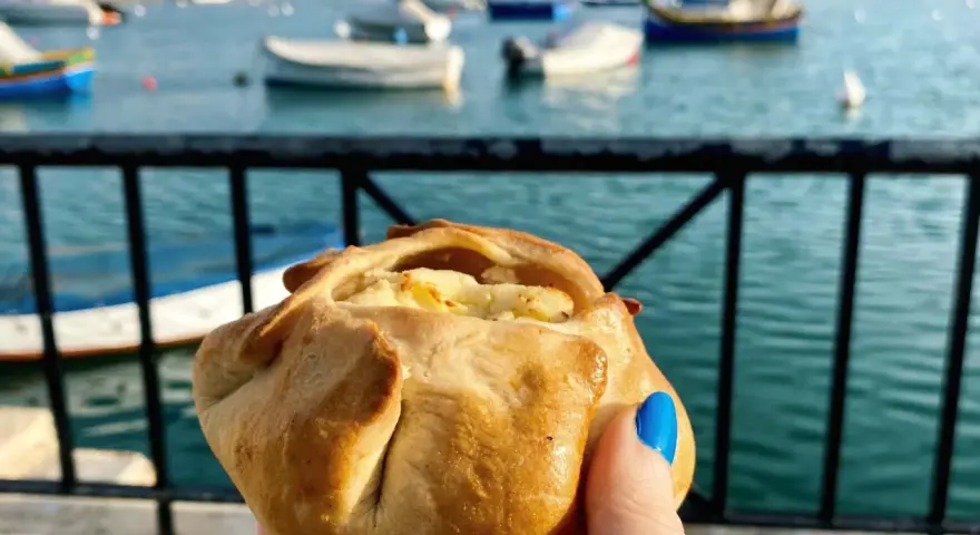 Ręka trzymająca pieczone ciastko z serem na tle maltańskiego portu z łodziami.