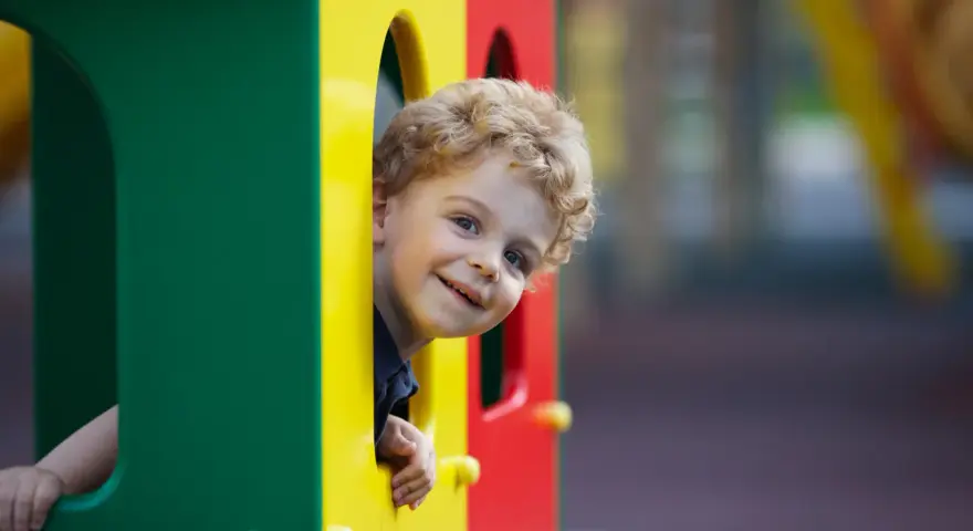 Uśmiechnięty chłopiec z blond lokami wygląda z kolorowego placu zabaw.