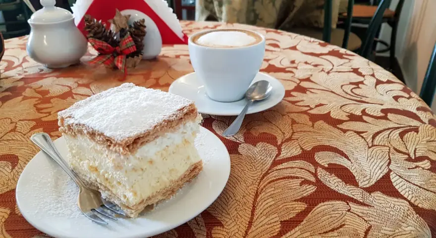 Kremówka posypana cukrem pudrem i filiżanka kawy na stoliku.