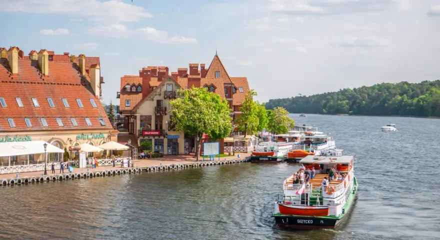 Port w Mikołajkach z zacumowanymi statkami wycieczkowymi i budynkami z czerwonymi dachami. Wspaniałe atrakcje w Mikołajkach czekają na turystów.