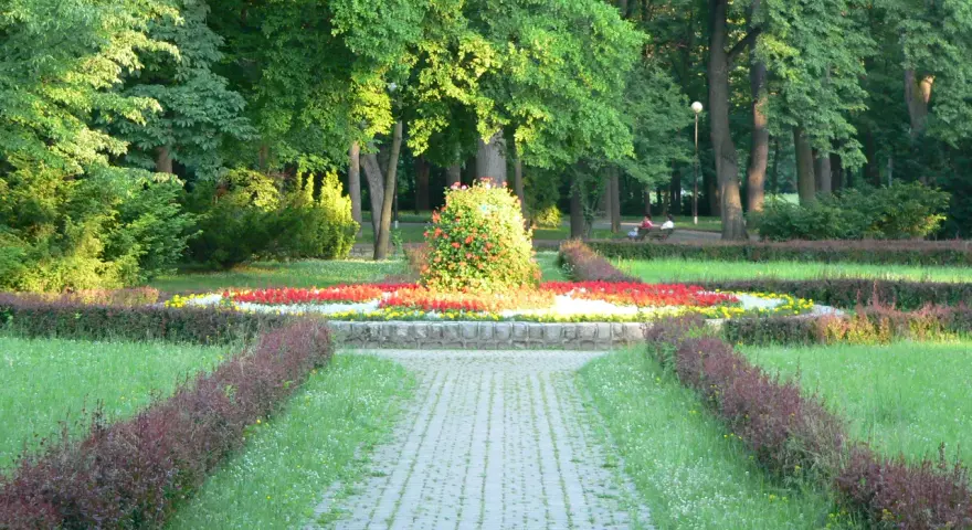 Kamienista ścieżka prowadzi przez park z kwitnącymi rabatami kwiatowymi i zielenią.