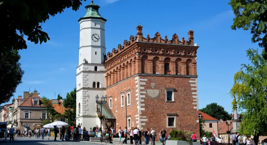 Ratusz w Sandomierzu z zegarem i zegarem słonecznym, na tle błękitnego nieba i drzew.