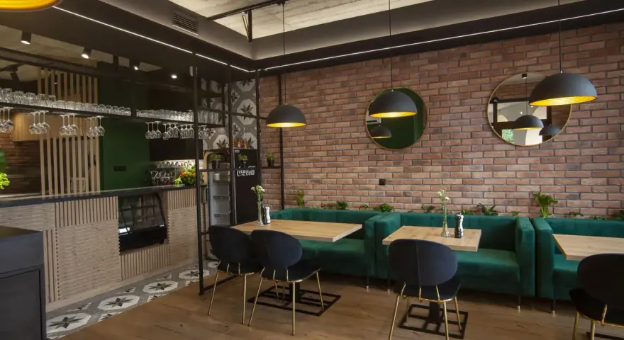 Nowoczesne wnętrze restauracji z ceglaną ścianą, zielonymi kanapami i drewnianymi stołami.
