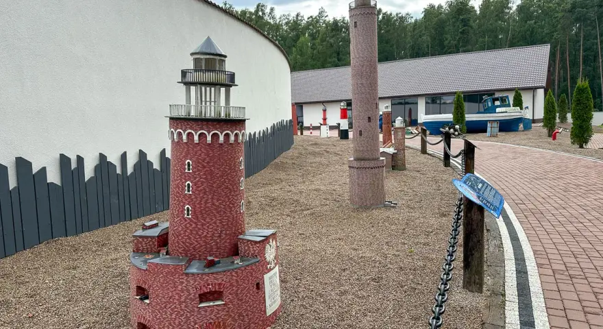 Miniatury latarni morskich na żwirowym placu, z budynkiem i łodzią w tle.