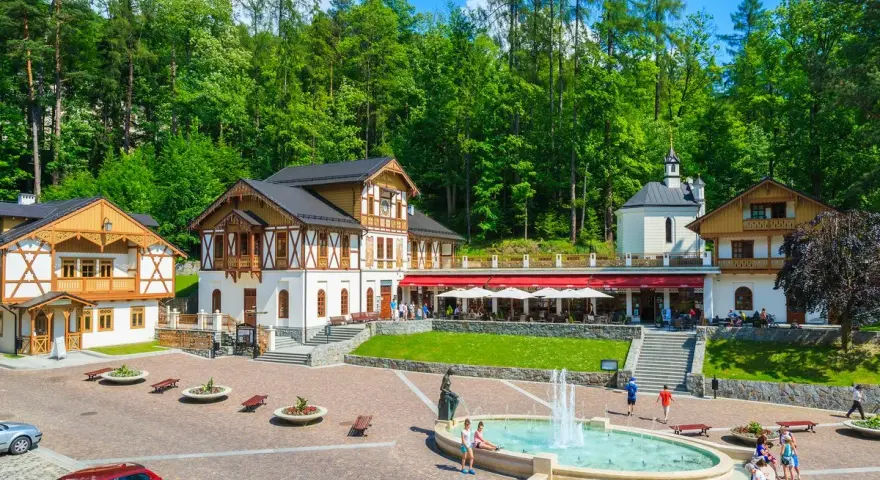 Plac Dietla w Szczawnicy z fontanną, budynkami uzdrowiskowymi i lasem w tle.