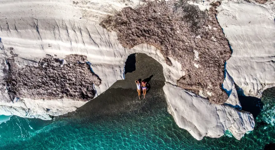 Dwie osoby leżą na czarnej plaży pod skalnym łukiem, obok turkusowej wody.