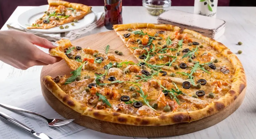 Pizza z krewetkami, oliwkami i kaparami na drewnianej desce, ręka podnosi kawałek.