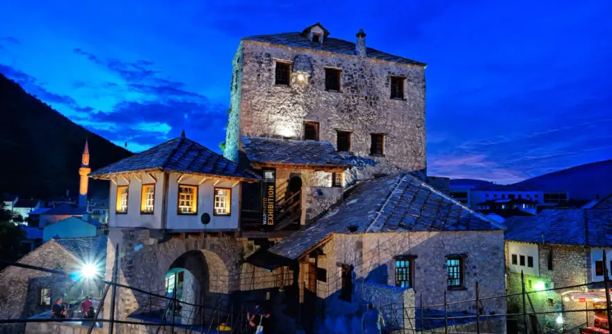 Kamienne budynki i most w Mostarze o zmierzchu, z widocznym minaretem w tle.