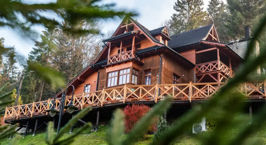 Drewniany góralski pensjonat z tarasem i balustradą, otoczony drzewami.
